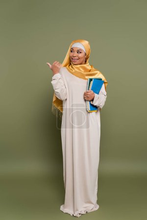 Smiling multicultural student in hijab holding notebooks and showing thumb up on green background