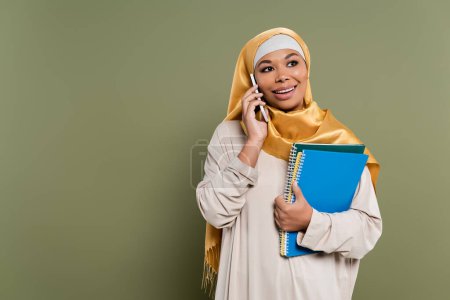 Cheerful multiracial student in hijab talking on smartphone and holding notebooks on green background