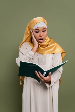 Shocked multiracial student in hijab holding notebook on green background