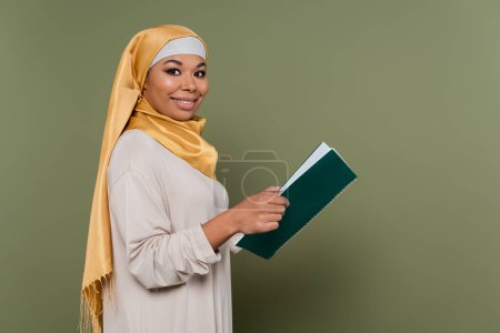 Positive multicultural student in hijab holding notebook and looking at camera on green background