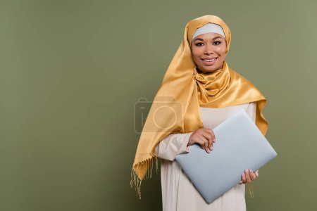 Positive multirassische Frau im Hijab hält Laptop in der Hand und schaut in die Kamera auf grünem Hintergrund