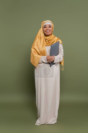Carefree multiracial woman in hijab holding laptop on green background