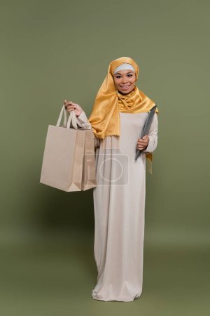 Multikulti-Frau im Hijab mit Laptop und Einkaufstaschen auf grünem Hintergrund
