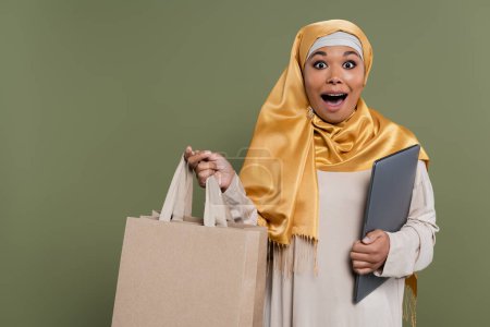 Aufgeregte multikulturelle Frau im Hidschab mit Laptop und Einkaufstaschen in der Hand 