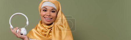 Carefree multiracial woman in hijab holding wireless headphones isolated on green, banner 