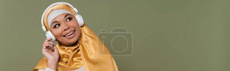 Fröhliche Frau im Hijab, die mit Kopfhörern Musik hört, isoliert auf grünem Banner 