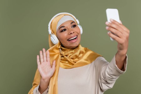Cheerful multiracial woman in hijab and headphones having video call on smartphone isolated on green 