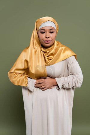 Multiracial woman in hijab feeling stomach pain on green background