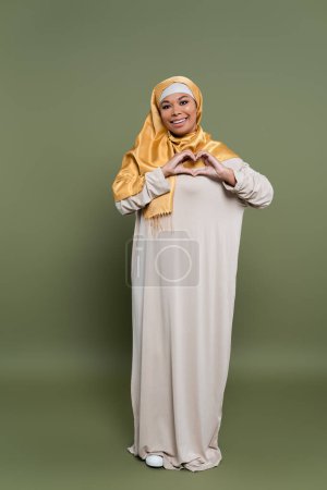 Téléchargez les photos : Femme multiraciale insouciante dans le hijab montrant signe d'amour avec les mains sur fond vert - en image libre de droit