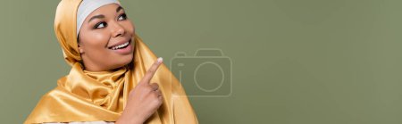 Smiling multiracial woman in hijab pointing with finger isolated on green, banner 