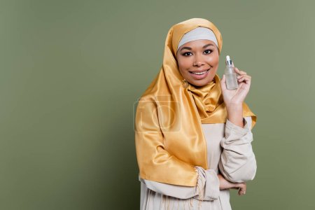 Positive multiracial woman in hijab holding serum on green background