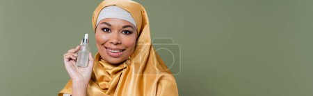 Multiracial woman in golden hijab holding serum isolated on green, banner 
