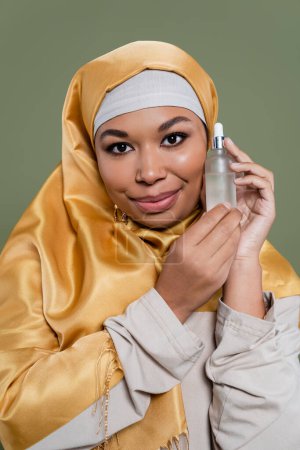 Portrait of multiracial woman in hijab holding cosmetic serum isolated on green 