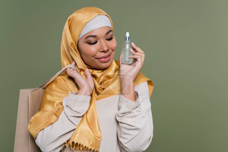 Smiling multiracial woman in hijab holding shopping bag and serum isolated on green 