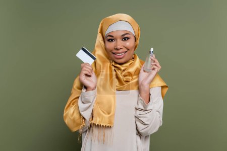 Smiling multiracial woman in hijab holding credit card and serum on green background