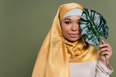 Joven mujer multirracial en hijab sosteniendo hoja tropical aislada en verde 