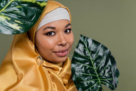 Porträt einer multirassischen Frau im Hijab, die in die Kamera schaut, in der Nähe von Blättern, die auf Grün isoliert sind 