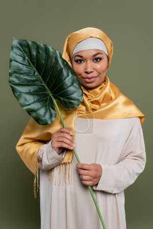 Joven mujer multirracial en hijab sosteniendo hoja tropical sobre fondo verde