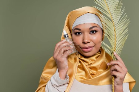 multiracial muslim woman in yellow satin hijab holding tropical leaf and bottle of cosmetic serum isolated on green