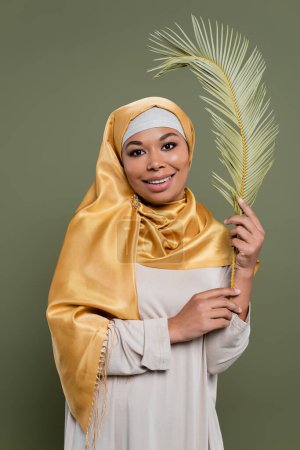 alegre mujer multirracial en amarillo hijab de seda sosteniendo hoja de planta exótica y sonriendo a la cámara aislada en verde