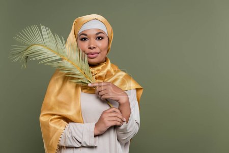 happy multiracial woman with makeup wearing yellow satin hijab and holding exotic leaf isolated on green