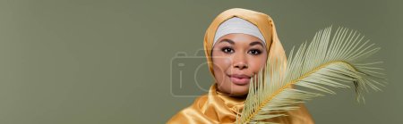 young multiracial woman in yellow silk hijab smiling at camera near leaf of tropical plant isolated on green, banner