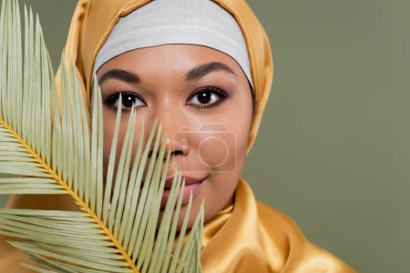 Porträt einer multirassischen muslimischen Frau mit Make-up, die in die Kamera in der Nähe von tropischen Blättern schaut, die auf Grün isoliert sind
