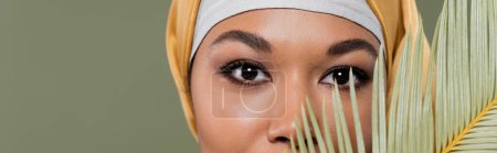 vista recortada de la mujer musulmana multirracial con maquillaje mirando a la cámara cerca de hoja exótica aislado en verde, pancarta