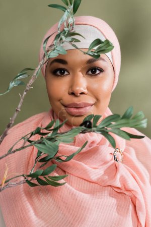 portrait of young multiracial woman in pink hijab smiling at camera near olive branch on green background