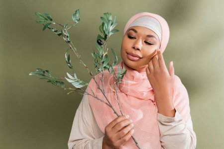 positive multiracial woman in pink hijab holding olive branch and touching perfect face on green background