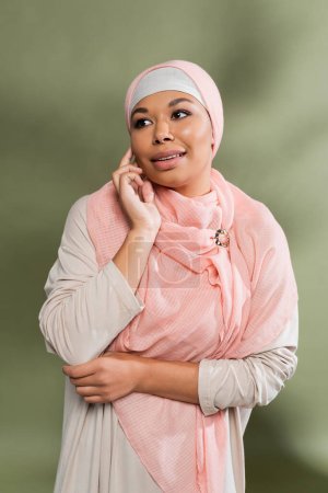 stylish multiracial woman in abaya dress and pink hijab touching face and looking away on green background