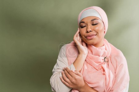 happy multiracial woman in pink hijab posing with closed eyes and touching perfect face on green background