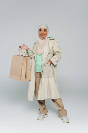 Photo pour Pleine longueur de femme multiraciale dans le hijab tenant des sacs à provisions tout en posant avec la main dans la poche de trench coat sur gris - image libre de droit