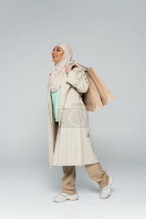 Photo for Full length of happy multiracial woman in hijab and stylish casual attire walking with shopping bags on grey background - Royalty Free Image