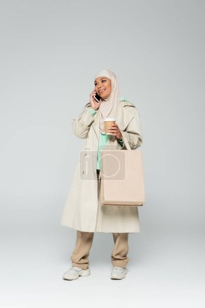Photo pour Pleine longueur de femme musulmane multiraciale joyeuse avec café à emporter et sacs à provisions parlant sur smartphone sur gris - image libre de droit