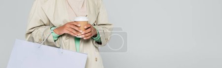 partial view of multiracial woman in trench coat holding shopping bag and disposable cup isolated on grey, banner