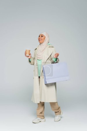 Photo pour Pleine longueur de femme multiraciale insouciante en tenue tendance et hijab tenant sac à provisions et boisson à emporter sur gris - image libre de droit