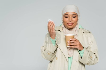 mujer joven multirracial en gabardina y hijab disfrutando del sabor del café aromático en una taza de papel aislada en gris