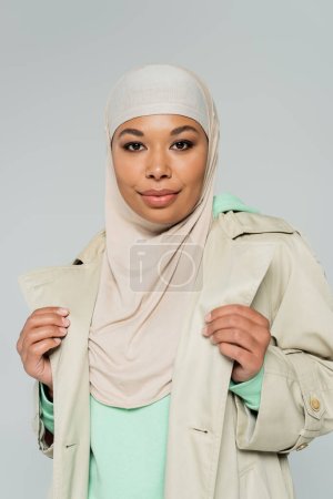Porträt einer stilvollen multirassischen Frau in Hijab und Trenchcoat, die isoliert auf grau in die Kamera blickt