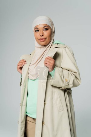 fashionable multiracial woman in hijab and stylish trench coat looking away and smiling isolated on grey