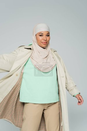 carefree multiracial woman in muslim hijab and trench coat posing and smiling at camera isolated on grey