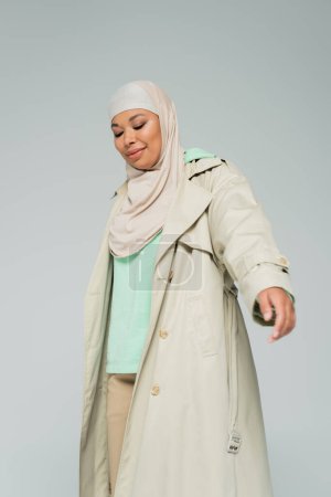 young and smiling multiracial woman in hijab and stylish trench coat posing isolated on grey