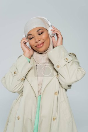 fröhliche und stilvolle multirassische muslimische Frau mit geschlossenen Augen, die Musik in drahtlosen Kopfhörern isoliert auf grau hört