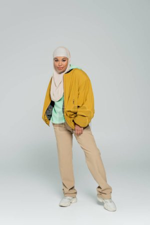 in voller Länge von multirassischen Frau im Hijab und trendigen lässigen Outfit stehend und lächelnd in die Kamera auf grauem Hintergrund