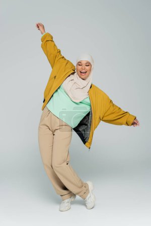 full length of multiracial muslim woman in yellow bomber jacket and beige pants posing with outstretched hands on grey background