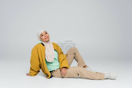full length of carefree multiracial woman in hijab and fashionable casual attire sitting on grey background