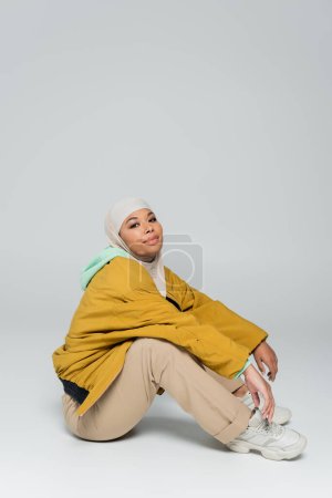 full length of pleased multiracial muslim woman sitting in beige pants and yellow jacket on grey background