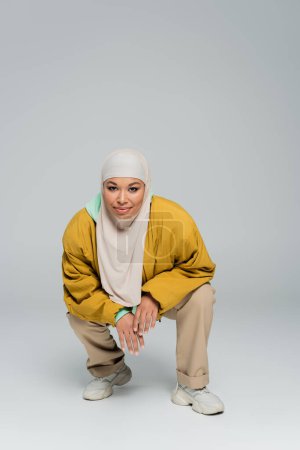 full length of fashionable multiracial woman in yellow bomber jacket and muslim hijab sitting on haunches on grey background