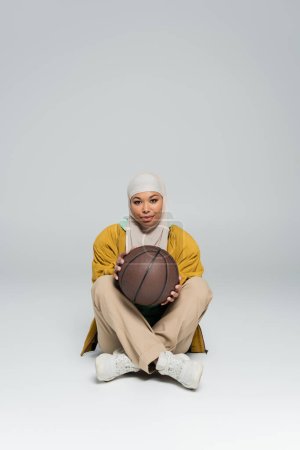 full length of smiling multiracial woman in hijab and pants with sneakers holding basketball while sitting on grey background