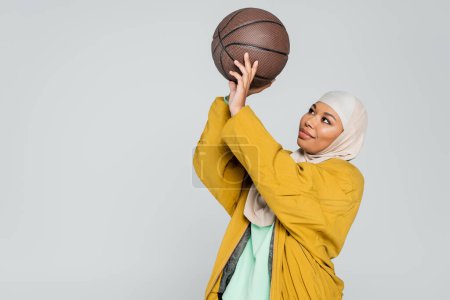 positive multiracial woman in hijab and bomber jacket playing basketball isolated on grey